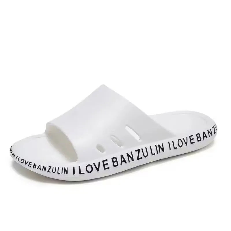 White unisex slippers with the text "I Love Banzulin" printed on them. The slippers are indoors on a wooden floor.