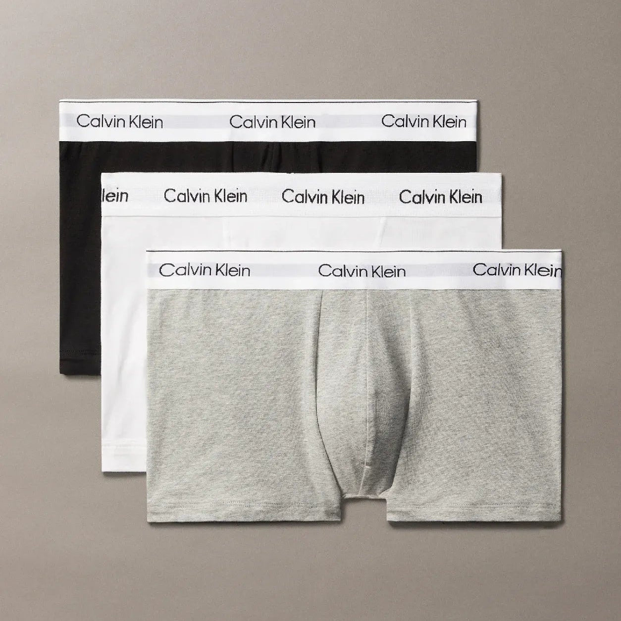 Three pairs of Calvin Klein boxer briefs in black, white, and gray on a beige background.