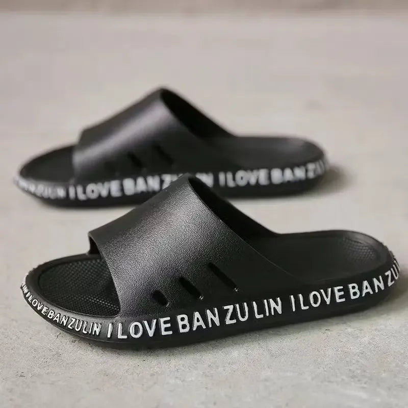 White unisex slippers with the text "I Love Banzulin" printed on them. The slippers are indoors on a wooden floor.