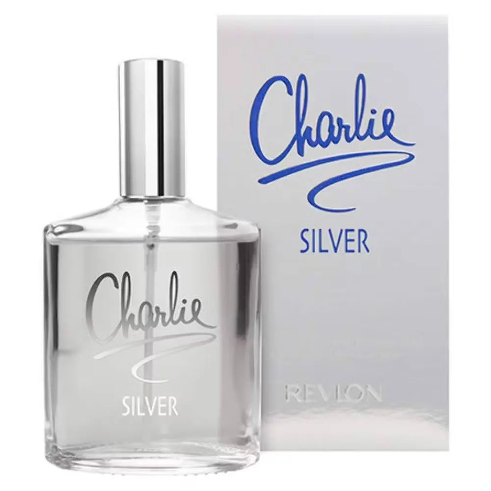 Revlon Charlie Silver Eau De Toilette bottle displayed on a mirrored vanity with soft lighting. Delicate white flowers and a silver pendant add a touch of sophistication.