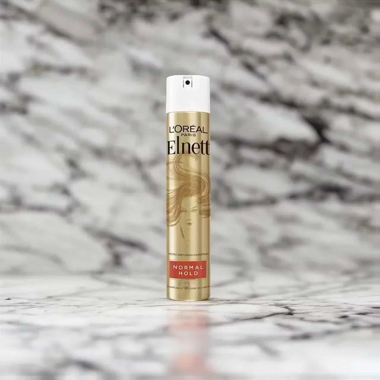 Silver can of L'Oreal Paris Elnett Micro Hairspray (250ml) with a pink nozzle and the text "Normal Hold with Precision Mist." A hand is spraying the hairspray onto styled hair.
