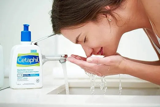 Large white bottle with blue pump dispenser labeled "Cetaphil Gentle Skin Cleanser 500ml." A hand pumps the dispenser, dispensing white cleanser onto another hand.
