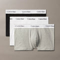 Three pairs of Calvin Klein boxer briefs in black, white, and gray on a beige background.